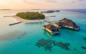 Overwater villas connected by wooden walkways sit above clear turquoise water hotel near a small tropical island with white sand beaches and palm trees.